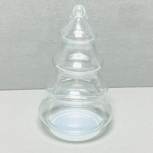 Puede incluir: Un recipiente transparente en forma de árbol de Navidad de vidrio. El árbol tiene un diseño escalonado con una base redondeada y una parte superior puntiaguda. El vidrio es transparente, lo que permite el paso de la luz. El recipiente es probablemente para fines decorativos.