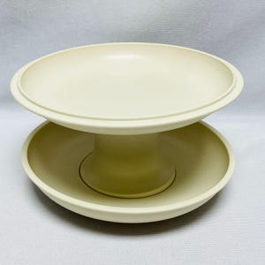 Puede incluir: Un soporte de servicio de dos niveles en color crema, con una base central de pedestal que conecta dos cuencos circulares poco profundos. La estructura está hecha de un material sólido y liso con un acabado mate uniforme en todo el objeto.
