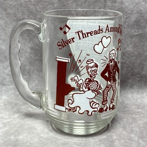 Puede incluir: Una taza de vidrio transparente con un diseño rojo que presenta dos figuras de dibujos animados, una mujer tocando un piano y un hombre de pie a su lado. El texto "Silver Threads Among the" es visible en la taza.