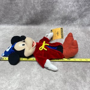 Puede incluir: Un peluche de Mickey Mouse, tumbado boca arriba, con un abrigo de terciopelo rojo, orejas negras y un sombrero azul y blanco. El juguete tiene guantes y zapatos blancos. Una etiqueta amarilla est&aacute; adjunta al juguete.