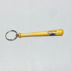 May include: A light brown wooden baseball bat keychain with a silver metal ring. The bat features the blue printed text TRI-CITY POSSE and a graphic logo of a baseball player silhouette integrated into the letters on the barrel of the bat.