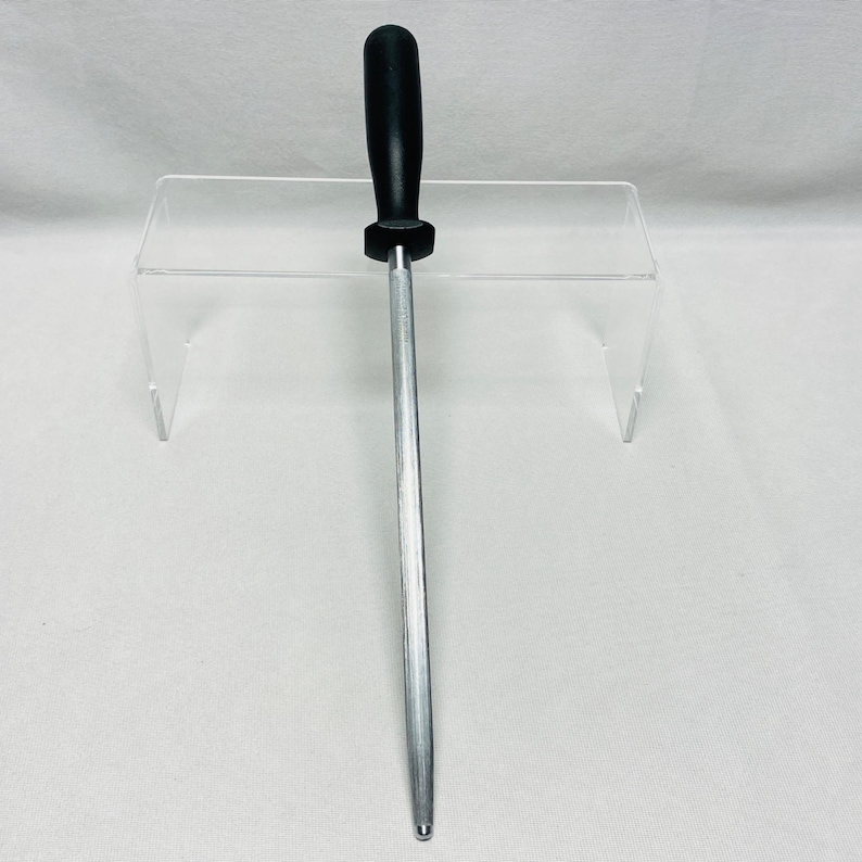 May include: A silver steel knife sharpening rod with a black handle. The rod is long and cylindrical, designed for honing the edge of knives. The handle is made of a smooth, solid material.