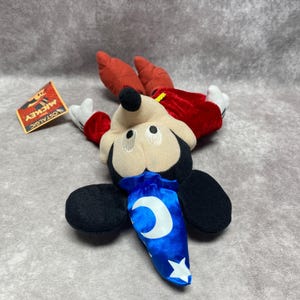 Puede incluir: Un peluche de Mickey Mouse vestido de hechicero, con capa y sombrero rojos. El peluche tiene orejas negras, cara beige y una bolsa azul con dise&ntilde;o de luna creciente y estrella. Una etiqueta dice "NOSTALGIC MICKEY".