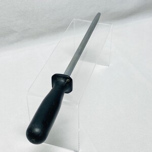 May include: A steel knife sharpening rod with a black handle. The rod is tapered and has a smooth, gray surface. The handle is made of a solid, black material and is ergonomically shaped for a comfortable grip. The sharpening rod is designed for kitchen use.