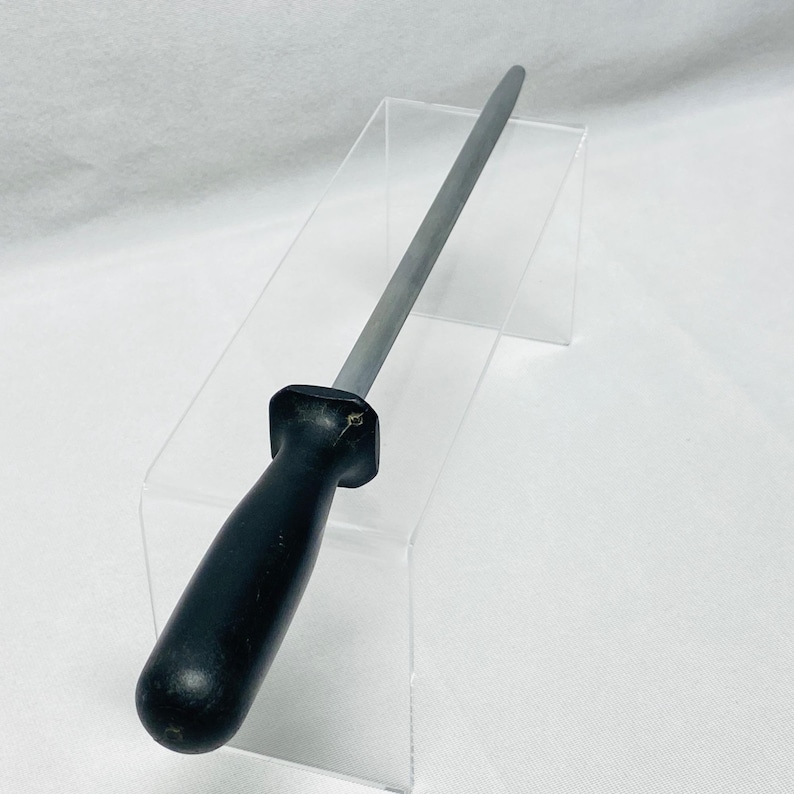 May include: A steel knife sharpening rod with a black handle. The rod is tapered and has a smooth, gray surface. The handle is made of a solid, black material and is ergonomically shaped for a comfortable grip. The sharpening rod is designed for kitchen use.