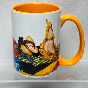 May include: White ceramic mug with an orange interior and handle. The mug features a colorful illustration of a person reclining on a patterned surface. The artwork includes red, blue, yellow, and black geometric shapes.