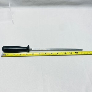 May include: A black-handled sharpening steel, approximately 13 inches long, with a silver-colored steel rod. A yellow measuring tape is placed next to the steel, showing the length. The handle is rounded and ergonomic.