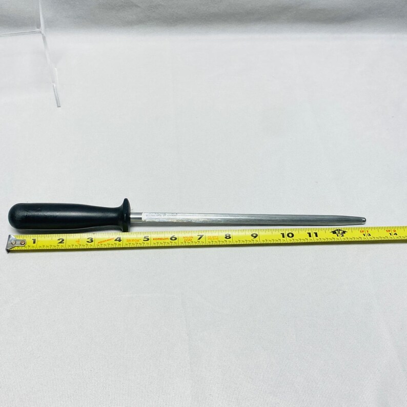 May include: A black-handled sharpening steel, approximately 13 inches long, with a silver-colored steel rod. A yellow measuring tape is placed next to the steel, showing the length. The handle is rounded and ergonomic.