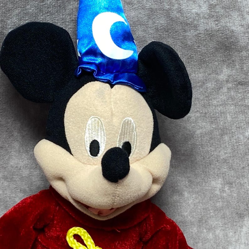 Puede incluir: Un peluche de Mickey Mouse disfrazado de hechicero. Mickey tiene orejas negras, una cara color crema y una t&uacute;nica roja. Lleva un sombrero de mago azul con una luna creciente blanca. El juguete es suave y detallado.