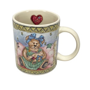 May include: A ceramic coffee mug with a colorful illustration of a teddy bear wearing a blue bow and holding a smaller teddy bear. The mug has a patchwork design with a red heart and safety pin on the top.