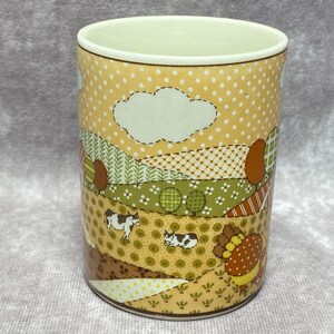 May include: A ceramic mug with a patchwork design featuring a brown and white cow, a brown and white sleeping cow, and a brown and orange turkey. The background is a yellow and brown patchwork design with a white cloud.