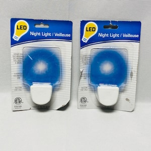 May include: Two LED night lights in packaging. Each night light has a blue, circular light and a white base. The packaging is blue and white, with the words "Night Light / Veilleuse" and "LED" printed on it.