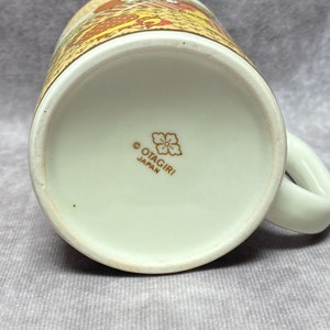 May include: The base of a white ceramic mug featuring a gold floral design and the inscription "OTAGIRI JAPAN".