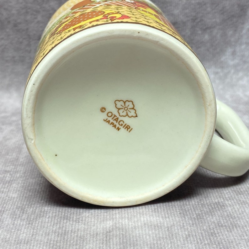 May include: The base of a white ceramic mug featuring a gold floral design and the inscription "OTAGIRI JAPAN".