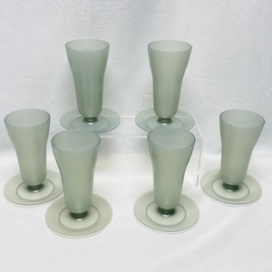Könnte beinhalten: Sechs Gläser aus mattgrünem Glas mit passenden runden Untertassen. Die Gläser haben eine ausgestellte, tulpenartige Form. Die Untertassen sind in einem helleren Grünton gehalten. Die Gläser sind auf einer weißen Oberfläche angeordnet.