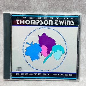 Puede incluir: Caja de CD del álbum "The Best of Thompson Twins". La portada presenta el nombre de la banda en blanco y un gráfico colorido de formas interconectadas en morado, rosa y azul. Los títulos de las canciones están listados.