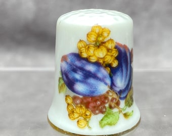 May include: A white porcelain thimble with a gold rim, decorated with a colorful fruit design. The design features two large purple plums, yellow grapes, and red grapes. The thimble has a textured top.