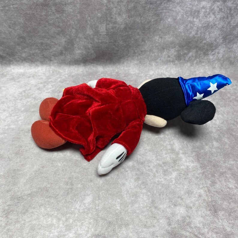 Puede incluir: Un peluche de Mickey Mouse disfrazado de hechicero, tumbado de lado. El juguete presenta una cabeza negra con orejas grandes, un sombrero de mago azul con estrellas blancas, una t&uacute;nica roja de terciopelo y guantes blancos con rayas negras.