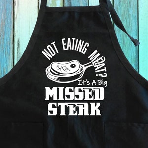 May include: Black kitchen apron with a white graphic that says "Not eating meat? It's a big missed steak." The graphic includes a cartoon image of a steak in a frying pan.
