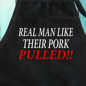 May include: Black apron with white text that reads "REAL MAN LIKE THEIR PORK PULLED!!" in a bold, sans-serif font. The word "PULLED!!" is in red. The apron is designed for cooking and grilling.