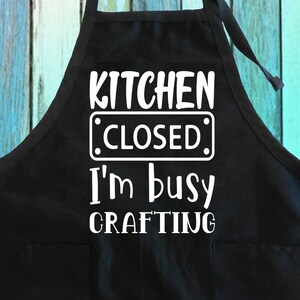 May include: Black kitchen apron with white text that reads "Kitchen Closed I'm Busy Crafting".
