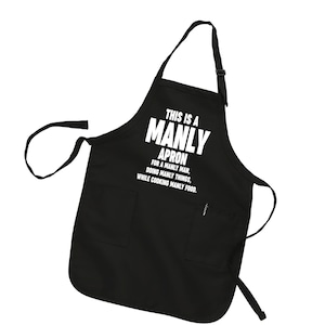 Puede incluir: Delantal negro con un bolsillo blanco y el texto "This is a Manly Apron for a Manly Man, Doing Manly Things, While Cooking Manly Food."