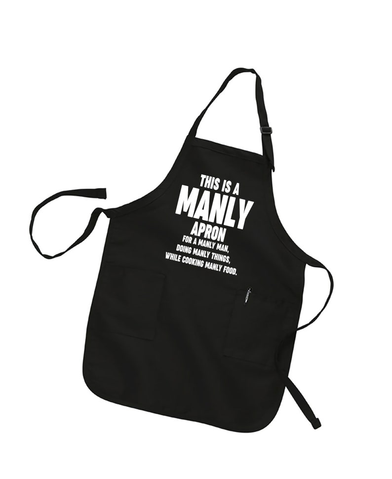 Puede incluir: Delantal negro con un bolsillo blanco y el texto "This is a Manly Apron for a Manly Man, Doing Manly Things, While Cooking Manly Food."