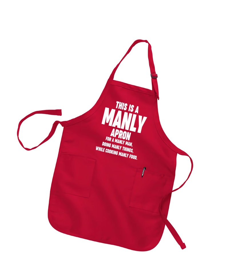 Puede incluir: Delantal rojo con un bolsillo blanco y el texto "This is a Manly Apron for a Manly Man, Doing Manly Things, While Cooking Manly Food."