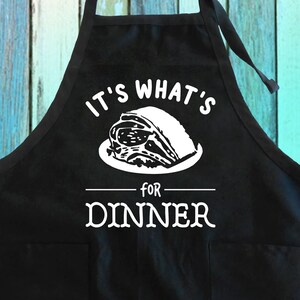 May include: Black apron with white text that reads "It's what's for dinner" and a graphic of a roast on a plate.