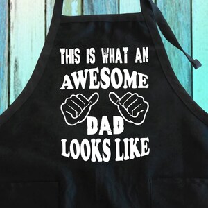 May include: Black apron with white text that reads "This is what an awesome dad looks like" with two thumbs up symbols.
