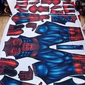 May include: A white fabric laid out with cut pieces of a Spider-Man costume. The costume design features a red and black web pattern on the arms, hands, and head, and a blue textured pattern on the torso and legs. The pieces are arranged on a white surface.