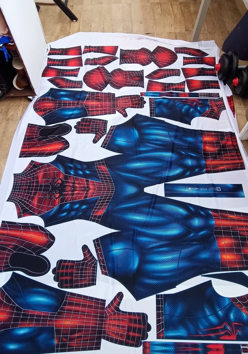 May include: A white fabric laid out with cut pieces of a Spider-Man costume. The costume design features a red and black web pattern on the arms, hands, and head, and a blue textured pattern on the torso and legs. The pieces are arranged on a white surface.
