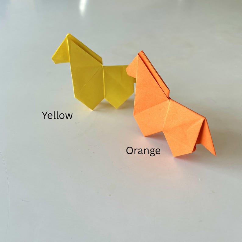 May include: Two origami horses, one yellow and one orange, are displayed on a white surface. The yellow horse is on the left, and the orange horse is on the right. The word "Yellow" is written below the yellow horse, and the word "Orange" is written below the orange horse.