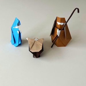 Puede incluir: Escena de la Natividad de origami con una figura azul, una figura marrón con un bastón y una figura marrón y beige. Las figuras son de papel plegado, creando una representación minimalista de la escena de la Natividad. Fondo neutro.