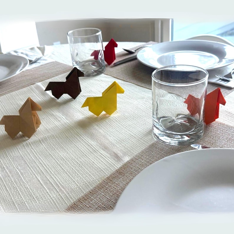 May include: A table setting with origami horse figures in brown, yellow, and red. Clear drinking glasses and white plates are arranged on a beige and white patterned tablecloth, creating a decorative and artistic display.