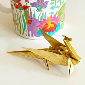 May include: A gold origami dragon with its wings spread out. The dragon is sitting on a white surface.
