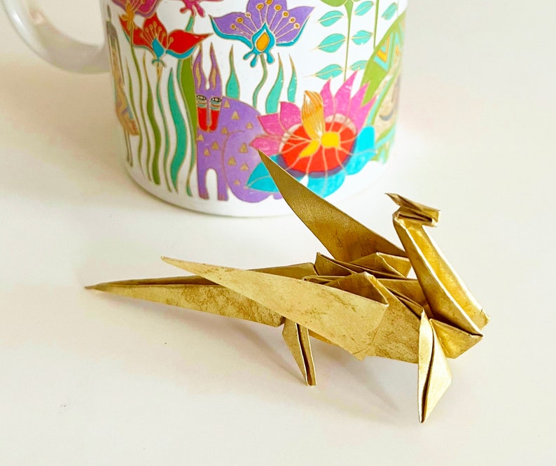 May include: A gold origami dragon with its wings spread out. The dragon is sitting on a white surface.