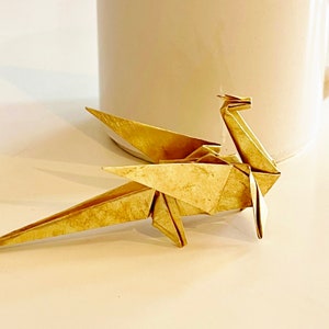 May include: A gold origami dragon, folded from paper, sits on a white surface. The dragon has a long tail and wings, and its head is tilted up.