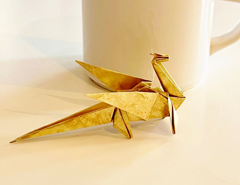 May include: A gold origami dragon, folded from paper, sits on a white surface. The dragon has a long tail and wings, and its head is tilted up.
