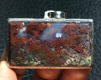 May include: A rectangular pendant with a silver frame and a colorful agate stone inside. The stone has a variety of brown, red, and white colors and patterns.