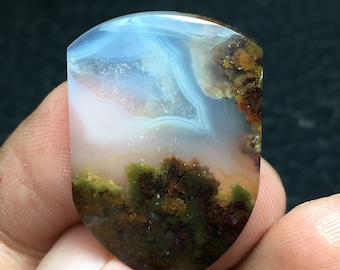 Könnte beinhalten: Ein polierter Achat-Cabochon mit einem einzigartigen Landschaftsmuster. Der Stein zeigt einen weißen und blauen Himmel mit braunen und grünen, moosartigen Formationen am Boden.