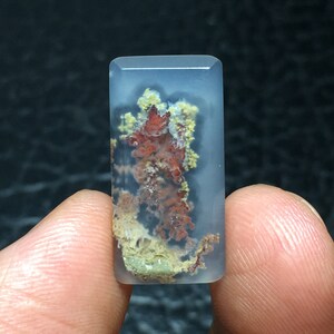 May include: A translucent, light blue agate cabochon with a dendritic pattern of red, yellow, and white. The agate is a rectangular shape and is approximately 1 inch in length.