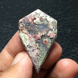 May include: A large, irregularly shaped, polished agate cabochon with a diamond-shaped outline. The stone has a variety of colors, including pink, red, gray, and white. The stone has a mottled, cloudy appearance.