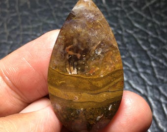 May include: A teardrop-shaped gemstone with a polished surface. The stone features layers of brown, gold, and white hues. The gemstone is held in a hand, showcasing its size and shape. The stone's natural patterns create a unique visual texture.