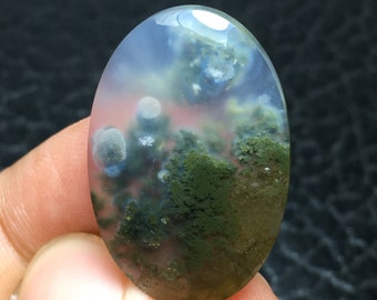 May include: Oval shaped green moss agate cabochon with white and gray inclusions. The stone has a translucent appearance with a cloudy, swirling pattern.