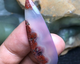 May include: A purple and white agate cabochon with red and brown dendritic inclusions. The cabochon is shaped like a teardrop and has a smooth, polished surface.