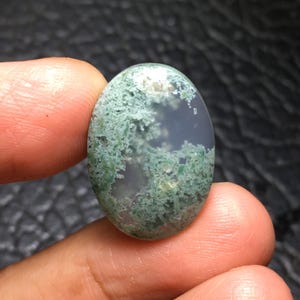 May include: Oval-shaped moss agate gemstone. The translucent stone features a landscape-like pattern of green moss-like inclusions within a clear, glassy base. The stone is held between two fingers.