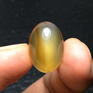 Könnte beinhalten: Ein polierter, oval geformter Edelstein mit transluzentem Aussehen. Der Stein weist eine Mischung aus goldgelben und braunen Farbtönen mit subtilen inneren Streifen auf. Die glatte Oberfläche reflektiert das Licht und erzeugt ein glänzendes Finish. Der Edelstein wird zwischen zwei Fingern gehalten.