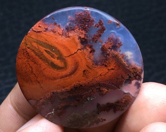 May include: A round, polished stone with a striking pattern of red, brown, and white hues. The stone's surface features intricate, swirling patterns and a unique, natural design. The stone is held in a hand against a dark background.