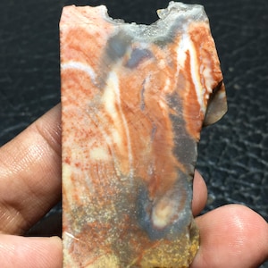 May include: A rough, natural stone with a unique pattern of red, white, and gray swirls. The stone has a jagged edge and a rough texture.